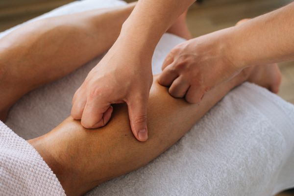 Close-up top view hands of unrecognizable male masseur with strong hands massaging lower leg of muscular athlete man after physical sports workout, lying on stomach at massage table.